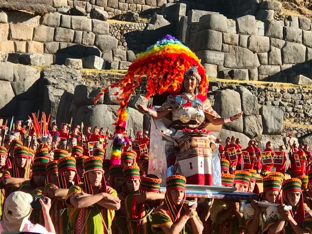 The Incan Queen arrives on a colorful throne-litter wearing gleaming silver discs and protected by a parasol of rainbow colors