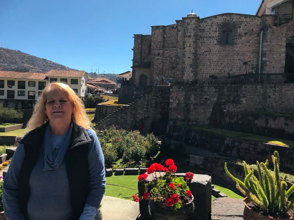 The author, Mona Rain, with blonde hair and a soft blue sweater, smiles with eyes closed as she is embraced by a sunbeam outside of Qoricancha, the ancient stone Temple of the Sun in Cusco, Peru 