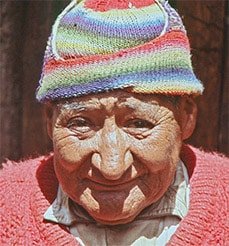 Don Benito Corihuaman Vargas appears in color in this photo, wearing what seems to be a red sweater, but bright pink in the direct sunlight. His knitted hat is quite worn, colorful against his weathered, bright face, with stripes of faded red, blue, purple, green, and white. He has a kind and gentle face.
