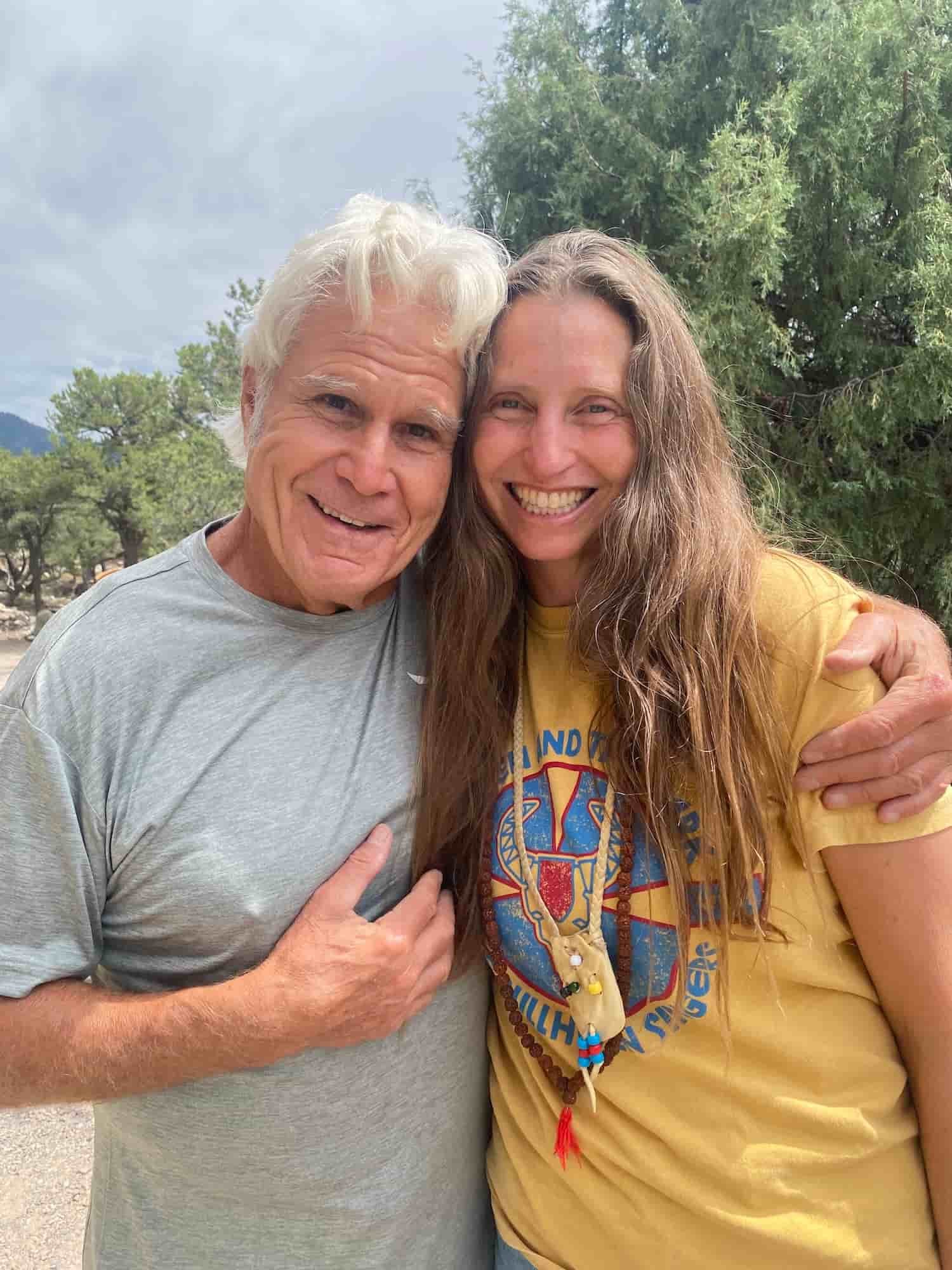 Don Oscar Miro-Quesada, smiling, white-haired and wearing a gray shirt embraces Naty Howard, a woman with long honey-colored hair and a yellow t-shirt, as they stand illuminated by the sun in Peru, posing for our Summer Solstice