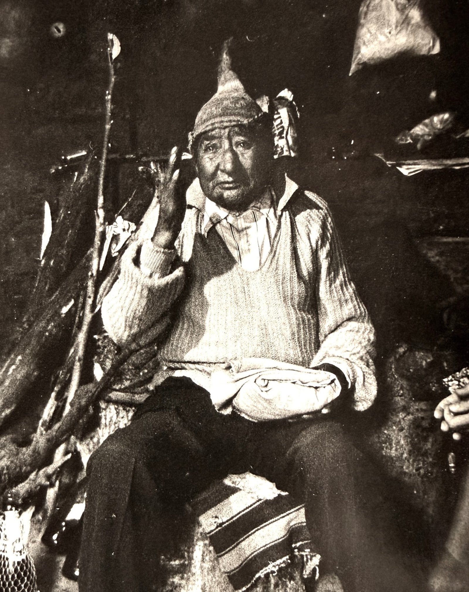 Don Benito sits on his worn, striped manta in preparation for a divination, holding his hand up mid-thought. He wears a thread-worn knitted cap and sweater in this black-and-white image.