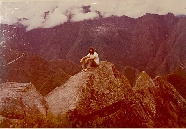 Old photo with faded color, dated in the 1980s, of a young man in a white t-shirt and wide-bottom pants, long hair and bearded, seated on a rocky outcropping with his arms resting on his knees. Behind him, the mountains of Andean Peru are shrouded in clouds.
